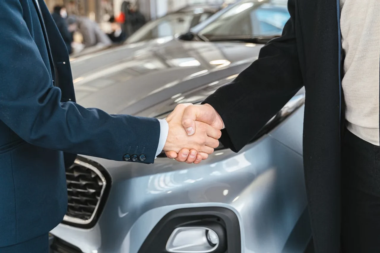 Poignée de main entre un vendeur et un client devant une voiture grise.